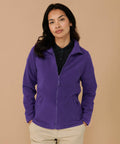 A person stands against a beige background wearing a purple Henbury HB851 zip-up fleece jacket and khaki pants. They have long dark hair and are looking into the camera with a neutral expression.