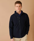 A person with short hair is wearing a Henbury HB860 navy zip-up jacket over a blue shirt and beige pants, standing against a beige background.