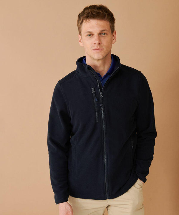 A person with short hair is wearing a Henbury HB860 navy zip-up jacket over a blue shirt and beige pants, standing against a beige background.