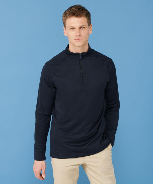A man in a long-sleeved, dark navy Henbury HB862 quarter-zip pullover stands against a light blue background with one hand in his beige pants pocket.