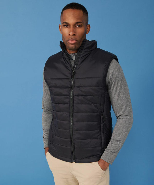 A person stands against a blue background, wearing a black Henbury HB875 quilted vest over a gray long-sleeve shirt and beige pants.