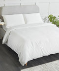 A neatly made bed with a grey upholstered HL001 headboard by Home & Living, white bedding, and two white pillows. A houseplant and a small bedside table are visible in the background.