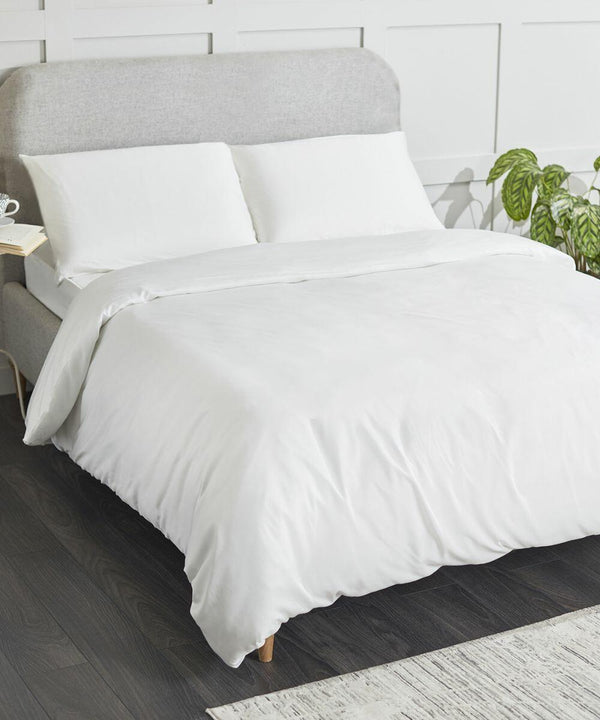A neatly made bed with a grey upholstered HL001 headboard by Home & Living, white bedding, and two white pillows. A houseplant and a small bedside table are visible in the background.