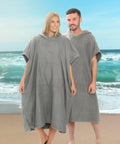 Two people stand on a beach wearing gray Home & Living HL211 hooded poncho towels, with the ocean and sky in the background.
