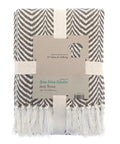 A folded brown and white herringbone-patterned blanket throw with white fringe is wrapped in cream bands. The label says "Green Living Collective Jazz Throw" and notes it’s made from 57 recycled clothing items.