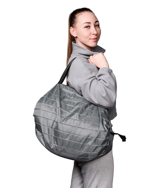 A woman in a light grey fleece jacket and tracksuit bottoms stands in profile, carrying the Home & Living Magic holdall/shopping bag—a large, round, grey quilted bag—over her shoulder against a white background.