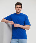 A man in a Russell J010M blue t-shirt and light blue jeans leans against a white wall, smiling at the camera.