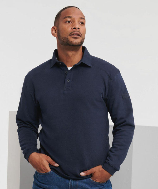 A man is wearing a dark blue long-sleeve Russell J012M polo shirt and blue jeans, standing against a white and gray background.