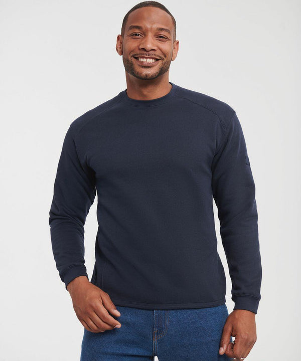 A man with a beard, wearing a navy blue Russell J013M long-sleeve shirt and blue jeans, smiles at the camera with one hand in his pocket.