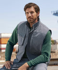 A man with a beard sits outdoors, wearing a green long-sleeve shirt and a gray sleeveless Russell J014M jacket. Industrial containers and equipment are visible in the background.