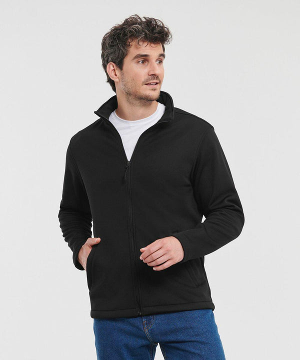 A man wearing a black Russell J040M zip-up jacket over a white t-shirt and blue jeans stands with his left hand in his pocket.
