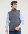 A man with curly hair is wearing a light blue shirt and a gray Russell J041M vest while looking to the side. He has his left hand partially in his pocket.
