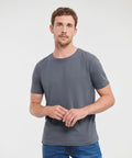 A man with short hair, wearing a gray Russell J108M t-shirt and blue jeans, stands against a plain white background.
