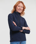 A person with red hair standing with arms crossed, wearing a navy blue Russell J140F jacket and blue jeans.