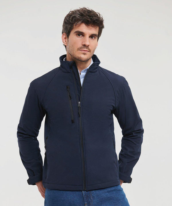 A man wearing a dark blue Russell J140M zip-up jacket over a light shirt stands with his hands in his pockets against a plain background.
