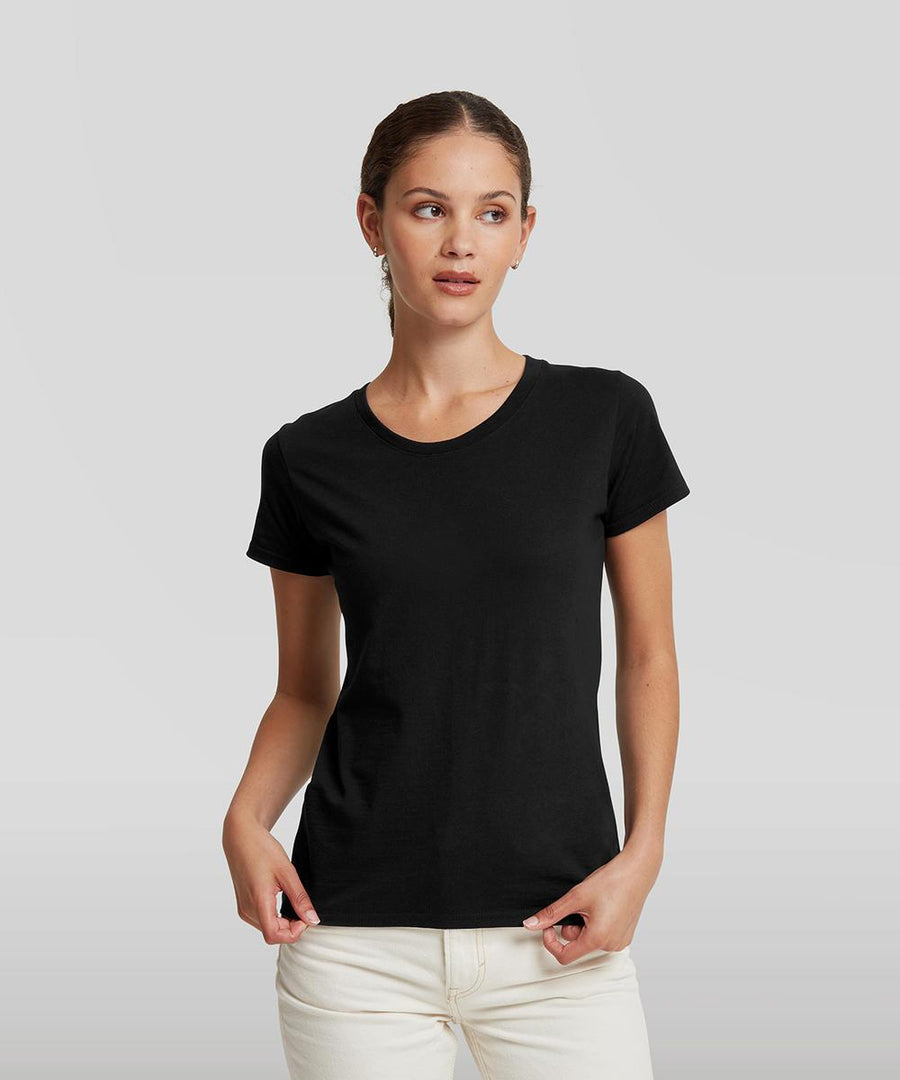 A woman stands against a plain grey background, wearing the Russell Athletic Women's authentic classic t-shirt in black and light-coloured trousers, looking slightly to the side with her hands in her pockets.