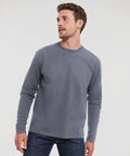 A man with short hair and a beard is wearing a grey long-sleeve J180L by Russell and jeans, looking to his left against a plain background.