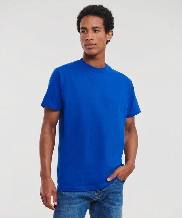A person stands wearing a plain blue J180M T-shirt by Russell and blue jeans against a plain background, with one hand in their pocket.
