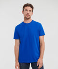 A man with short brown hair and a beard stands against a white background, wearing a plain blue J215M by Russell and dark jeans, looking at the camera.