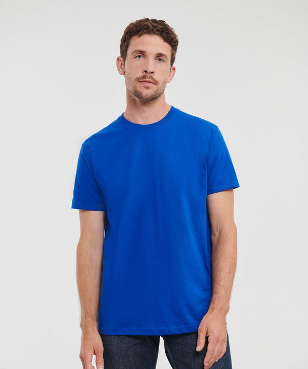 A man with short brown hair and a beard stands against a white background, wearing a plain blue J215M by Russell and dark jeans, looking at the camera.