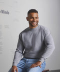 Man seated, wearing a grey Russell J262M sweatshirt and blue jeans, smiling and looking to his right. Minimalist background with faint text on the wall.