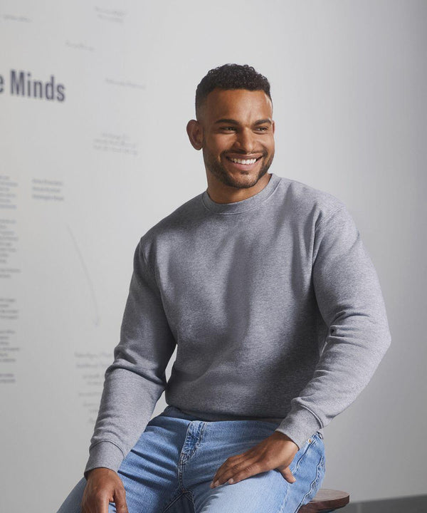 Man seated, wearing a grey Russell J262M sweatshirt and blue jeans, smiling and looking to his right. Minimalist background with faint text on the wall.
