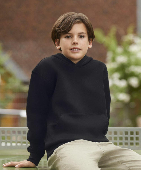A young boy with brown hair is wearing a black hoodie and beige pants. He is sitting outdoors on a bench with a blurred background of greenery and buildings, wearing the Russell J265B.