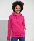 Person in a bright pink Russell J265F hoodie and jeans, smiling and leaning against a white surface.