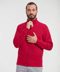 A man with a beard wearing a red Russell J267M jacket and gray pants stands against a plain background, holding the jacket collar with one hand and the zipper with the other.