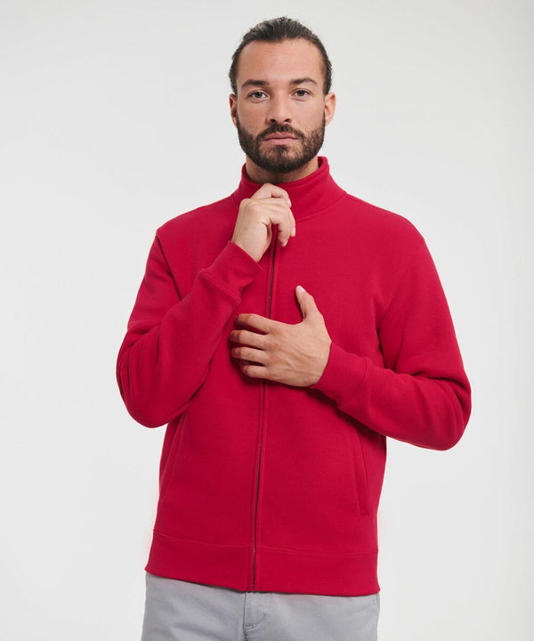 A man with a beard wearing a red Russell J267M jacket and gray pants stands against a plain background, holding the jacket collar with one hand and the zipper with the other.