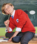 A child in a red sweater and glasses sits on a desk in front of a blackboard, holding a Russell J272B.