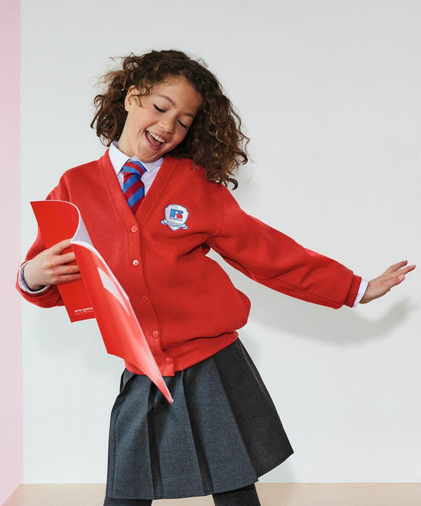 A young child wearing a red Russell school cardigan (J273B), white shirt, blue and red striped tie, and gray skirt stands holding a red folder, smiling and posing energetically.