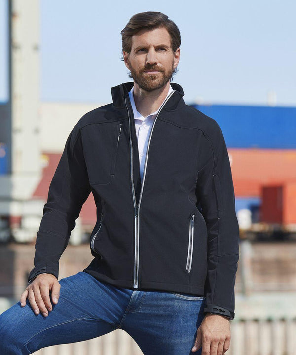 A man with short hair and a beard stands outdoors, wearing a black Russell J410M over a white shirt and blue jeans, with a backdrop of a building and colorful shipping containers.