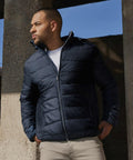 A man wearing a dark blue Russell J440M puffer jacket and beige pants stands outdoors, leaning against a concrete structure and looking to the left.