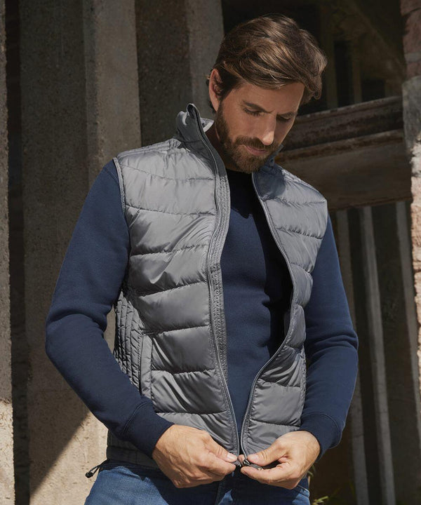 A man with a beard, wearing a navy blue long-sleeve shirt and a gray quilted vest, zips up the Russell J441M vest while standing in front of a concrete structure.