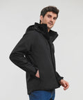 A man wearing a black Russell J510M hooded jacket stands against a plain background, with his left hand in the pocket and looking slightly to his left.