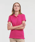 A person with long hair is wearing a bright pink J566F polo shirt by Russell and beige pants, standing with arms crossed against a plain white background.