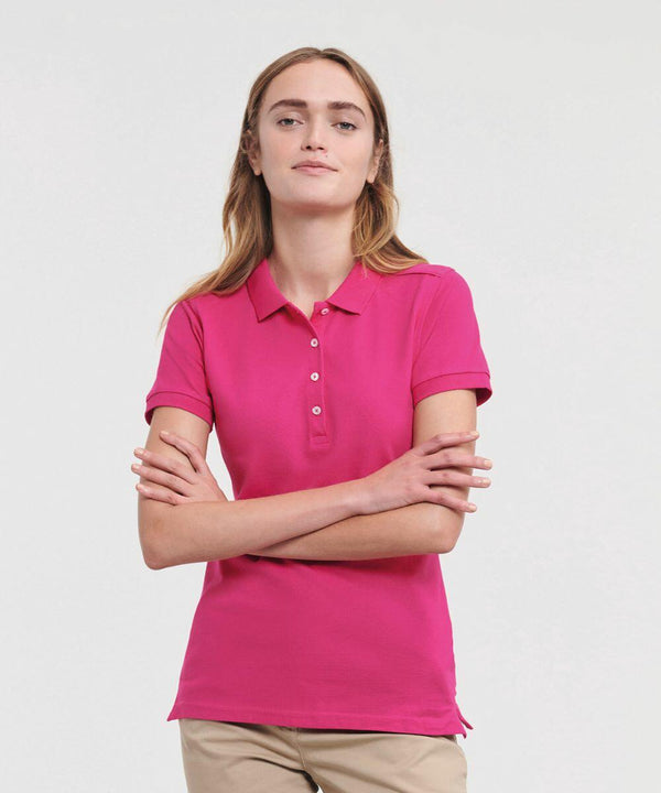 A person with long hair is wearing a bright pink J566F polo shirt by Russell and beige pants, standing with arms crossed against a plain white background.