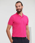 A man with a beard wearing a bright pink Russell J566M polo shirt and dark pants stands with his hands in his pockets, smiling.