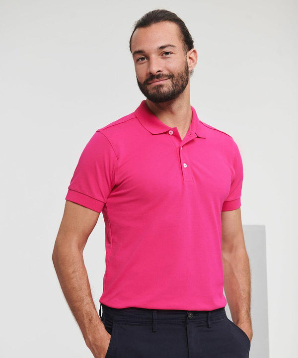 A man with a beard wearing a bright pink Russell J566M polo shirt and dark pants stands with his hands in his pockets, smiling.