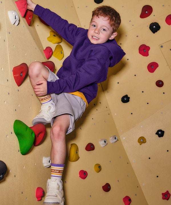 A young boy wearing a Russell J575B purple hoodie and gray shorts climbs an indoor rock wall, gripping colorful holds with a determined expression.