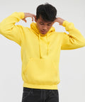 Person wearing a yellow Russell J575M hoodie with the hood down, standing against a plain background, adjusting the drawstrings while looking down.