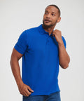 A man in a blue Russell J577M polo shirt adjusts his collar while looking to the side. He has short hair and is wearing blue jeans.