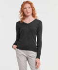 A person with red curly hair, wearing a dark gray long-sleeve Russell J710F V-neck sweater and light gray pants, stands against a plain white background, with one hand in their pocket.