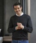 A man wearing a dark Russell J710M sweater and white shirt smiles and stands by a window with his hands clasped.