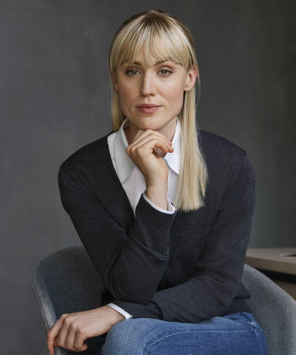 A woman with long blonde hair and bangs, dressed in a dark sweater over a white collared shirt, sits with her hand resting on her chin, wearing the Russell J715F.