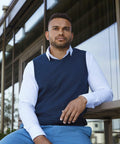 Man in a white shirt and navy Russell J716M sweater vest sits outside a glass building, looking away from the camera with a thoughtful expression.
