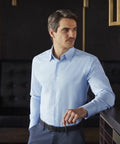 A man in a light blue Russell Collection J922M and dark pants stands indoors, leaning against a surface and looking off to the side.