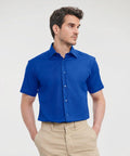 A person wearing a blue short-sleeved Russell J923M button-up shirt and beige pants stands against a plain background looking to the side.