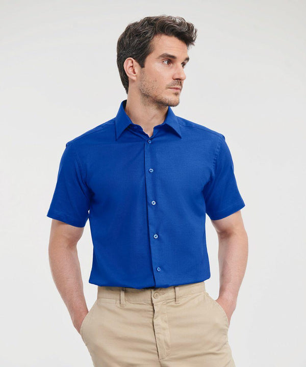 A person wearing a blue short-sleeved Russell J923M button-up shirt and beige pants stands against a plain background looking to the side.